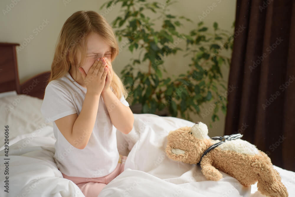 A little girl sits and sneezes on the bed among the blankets and pillows. House dust mite allergy