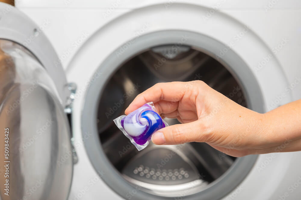Woman putting laundry detergent capsule into washing machine indoors, closeup.Colorful laundry eco gel in capsule. Washing clothes.The concept of washing and cleanliness