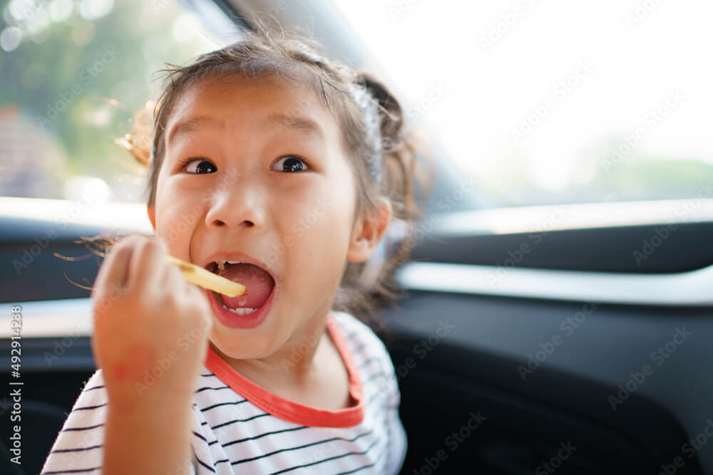 Little Asian young girl staying in the car and eating fried potato chips.